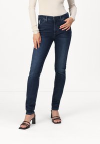 Sötétkék farmer skinny farmernadrág magas derékkal, öt zsebbel és enyhe kopott textúrával, fekete, pántos magassarkú szandálokkal párosítva.