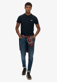 Camiseta de algodón azul marino, jeans ajustados azules, botas negras. Camisa de cuadros roja atada alrededor de la cintura para un toque adicional.