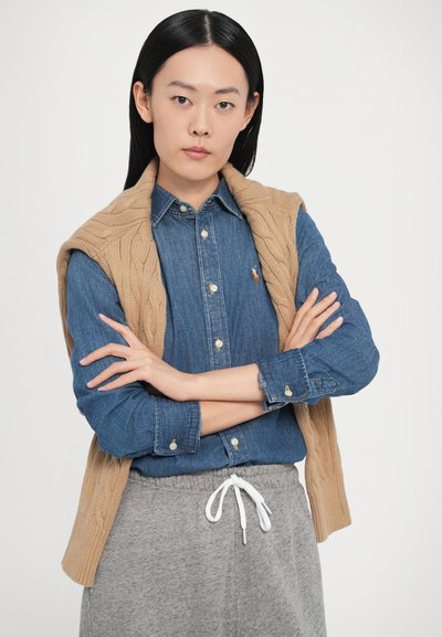 Pieskovohnedá vesta s pleteným vzorom cez modrú denimovú košeľu na zapínanie na gombíky. Sivé teplákové nohavice so šnúrkou v páse. Model stojí so založenými rukami pred neutrálnym pozadím.