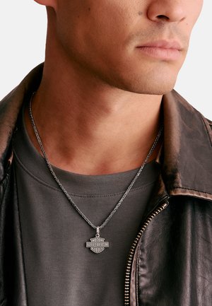 Nahaufnahme eines Mannes, der eine silberne Harley-Davidson-Anhänger-Halskette, ein graues Hemd und eine dunkelbraune Lederjacke trägt.