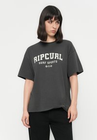 Kvinde med mørkt hår iført en oversized mørkegrå Rip Curl Surf Sports T-shirt og sorte bukser, stående foran en ensfarvet hvid baggrund.