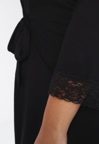 Abito nero con vita aderente e dettaglio annodato, tessuto morbido e accenti in pizzo sulle maniche per un'aggiunta di texture.