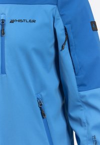 Modrá softshellová bunda s kapsami na zip, vysokým límcem a logem "Whistler". Látka má hladkou strukturu s voděodolným povrchem.