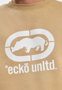 Sudadera beige con un gran gráfico blanco de un rinoceronte dentro de un logo circular y el texto "*eckō unltd." debajo. Material suave.
