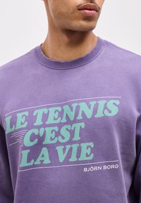 Άνδρας που φοράει μωβ φούτερ με πράσινο κείμενο «LE TENNIS C'EST LA VIE» και μικρότερο λευκό λογότυπο «BJÖRN BORG» σε απλό φόντο.
