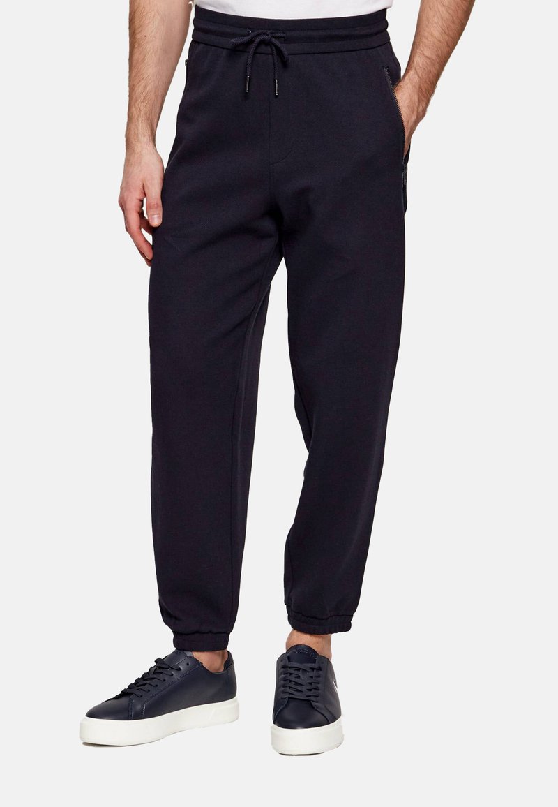 Homme portant un pantalon de jogging noir avec des poignets élastiques et des baskets noires à semelles blanches, debout devant un fond uni.