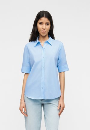 Ženska z dolgimi temnimi lasmi, oblečena v svetlo moder srajčni pulover s kratkimi rokavi in svetlo modre kavbojke, stoji na enostavni ozadju.