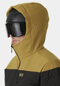 Skijacke mit einem gelb-schwarzen Farbblockdesign, ausgestattet mit einem Reißverschluss auf der Vorderseite, Kapuze und einem Logo auf der Brust. Glattes, strukturiertes Material.