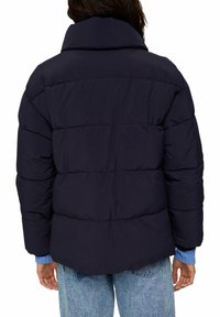 Person iført en mørkeblå puffjakke med bred krave og lyseblå sweatermanchetter synlige, parret med lyse denimjeans.