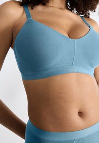 Lyseblå trådløs bh med en glat tekstur, der har tynde stropper og en v-udskæring. Sloggi-logoet er placeret på båndet nedenfor.