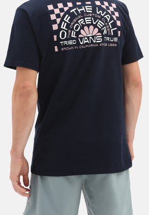Person iført en navyblå Vans T-shirt med pink ternet mønster og tekst på bagsiden, kombineret med lysegrå shorts.