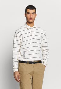 Langermet polo skjorte i krem med svarte horisontale striper, med krage og tekstdetalj på nederste venstre side. Kombinert med khaki bukser.
