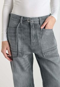 Pantalon en denim gris avec taille haute, plusieurs poches avant et une boucle de ceinture visible maintenue par un anneau en métal. Texture lisse.