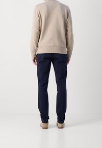 Persona in piedi con le spalle rivolte, che indossa un maglione beige, pantaloni blu scuro e scarpe beige su uno sfondo bianco.