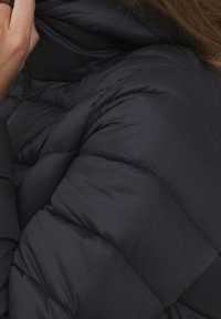 Veste matelassée bleu marine avec un design piqué et un col haut. Le matériau semble doux et isolé, avec une texture lisse.