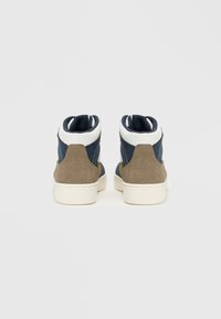 Korkeavartiset tennarit, joissa on tummansininen kangas ja vaaleanruskeat mokka yksityiskohdat. Valkoinen kuminääntö, jossa on kontrastoivaa ommelta. Pehmustettu nilkkacolari.