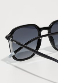 Ochelari de soare negri cu un cadru pătrat, lentile gri cu un gradient și brațe subțiri negre. Prezintă brandingul "HUGO" pe unul dintre brațe.