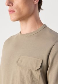 O sweatshirt beige de gola redonda apresenta um bolso em patch texturizado no lado esquerdo do peito, feito de um tecido leve e suave. Detalhe de gola canelada.