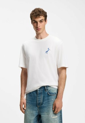 Junger Mann mit lockigem Haar, der ein weißes T-Shirt mit kleinem blauem Pferdemotiv und blaue, abgenutzte Jeans auf weißem Hintergrund trägt.