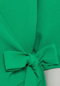 Zielony teksturowany strój z detalem wiązanym w talii, wykonany z pofałdowanej tkaniny o gładkim, lekko rozciągliwym wyglądzie.