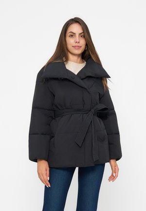 Kvinde iført en sort dunjakke med bælte og bred krave samt blå jeans, stående mod en ensfarvet hvid baggrund.