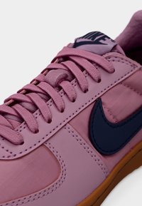 Adidași roz cu finisaj neted, având un logo albastru marin, șireturi texturate și talpă de cauciuc maro. Vedere de apropiere a secțiunii superioare.