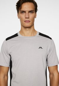 Grå idrottströja med svarta sidopaneler, korta ärmar, rund halsringning och subtilt logo på bröstet. Slät, andningsbar tyg.