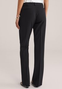 Kobieta ubrana w czarne spodnie o prostych nogawkach i biały top, pokazana od tyłu z ręką opartą na udzie i złotą bransoletką na nadgarstku.