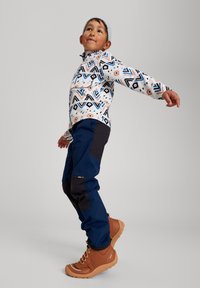 Bunte gemusterte Jacke mit geometrischen Designs, marineblaue Hose mit schwarzen Akzenten und braune wasserdichte Stiefel mit strukturiertem Sohlenprofil.