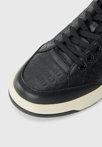 Tenis de piele neagră cu detalii în relief ale logo-ului, suprafață texturată și talpă albă contrastantă. Baticurile sunt negre, iar găurile pentru sfoară sunt metalice.