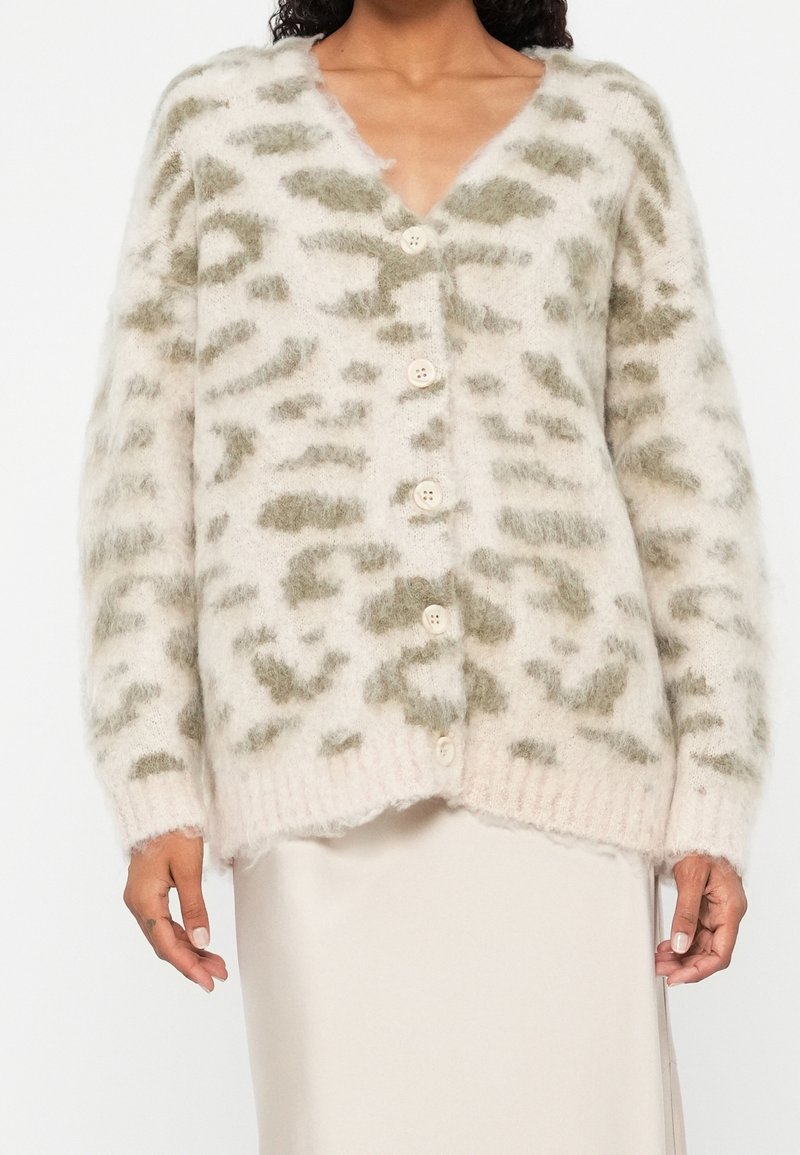 Cardigan duveteux blanc et gris à boutons avec un motif abstrait porté sur une jupe claire sur une personne.