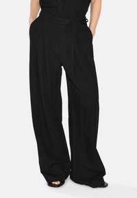 Pantalons larges noirs en tissu léger. Présentent une taille ajustée avec une ceinture nouée et des plis sur le devant pour un détail supplémentaire.