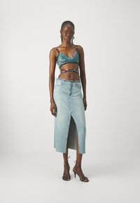 Un bralette turchese con motivo e spalline sottili, abbinato a una gonna di jeans chiari con spacco frontale e orlo sfrangiato. Sandali neri con tacco.