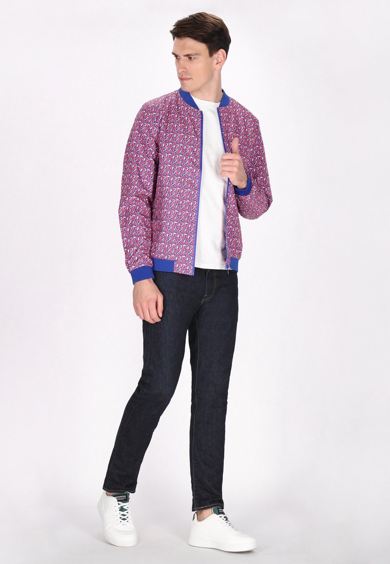 Giacca bomber rosa con un motivo geometrico, cuffie blu e chiusura con zip. Abbinata a jeans scuri e sneakers bianche.