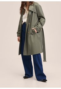 Trench-coat vert olive avec grand col, deux poches avant et ceinture. Associé à un jean bleu foncé à jambes larges et des chaussures noires simples.
