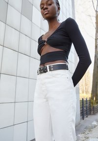 Camisola preta de mangas longas com um design recortado e detalhe em argola prateada, combinada com calças brancas de cintura alta e um cinto preto com ilhoses.