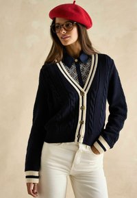 Jūras spēku adīts kardigāns ar kabeli, krēms akcentiem, lielām pogām, virs rakstainas krekliņas, kombinēts ar baltām biksēm un sarkanu bereti.