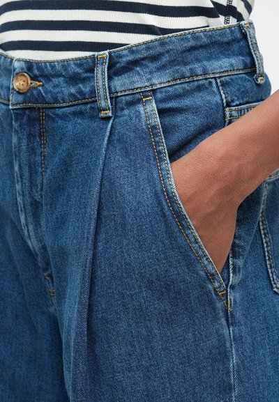 Hånd i høyre lomme på høy midje blå denimjeans, kombinert med en svart og hvit stripete skjorte.