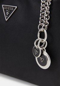Musta synteettinen käsilaukku, jossa on hopeinen ketjuhihna, kolmion muotoinen logo ja pyöreät koristemaalaukset teksturoidulla pinnalla.
