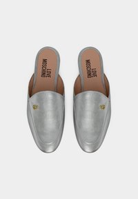 Ασημί μοκασίνια slip-on με μικρές χρυσές καρδιές και την επιγραφή "LOVE MOSCHINO" στις καφετί εσωτερικές σόλες, εμφανιζόμενα σε λευκό φόντο.