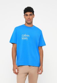 Mėlyna medvilninė marškinėliai su trumpomis Rankovėmis, apvaliu iškirpimu ir baltais užrašais "calvin klein." Paprastas dizainas, laisvas kirpimas, lygus tekstūra.