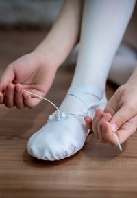Weiße Ballettschuhe aus dehnbarem Stoff, die mit einem dünnen Band geschnürt werden. Im Hintergrund ein heller Holzboden, Hände, die den Schuh anpassen.