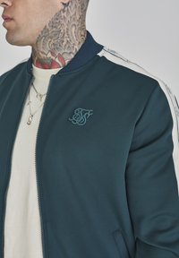 Chaqueta bomber verde para hombre con cuello acanalado, rayas laterales blancas y logotipo bordado. Fabricada en un tejido suave, cuenta con cierre de cremallera y bolsillos.