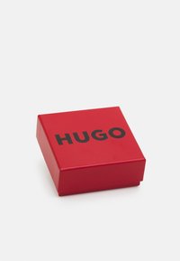 Boîte carrée rouge avec le texte noir "HUGO" sur le couvercle, placée sur un fond blanc.