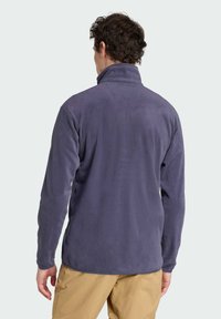 Fleece jacket in dark purple with a high collar, long sleeves, and a soft texture. The hem is slightly curved, worn over beige pants.