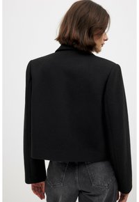 Chaqueta corta negra hecha de tejido texturizado, con un amplio cuello y hombros estructurados. La vista posterior muestra líneas limpias con un detalle mínimo.