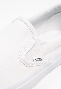 Bílé plátěné nazouvací boty s kulatou špičkou, strukturovaným svrškem a bílou gumovou podrážkou. Obsahují malou černou značku Vans.