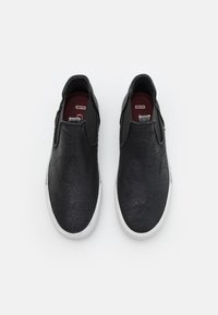 Chaussures noires à enfiler en matériau texturé, avec un bout rond, des panneaux latéraux élastiques et une semelle en caoutchouc blanche.