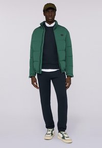 Giacca imbottita verde, maglione blu navy, camicia bianca e pantaloni della tuta scuri. Presenta trame lisce; le scarpe hanno accenti verdi e beige.