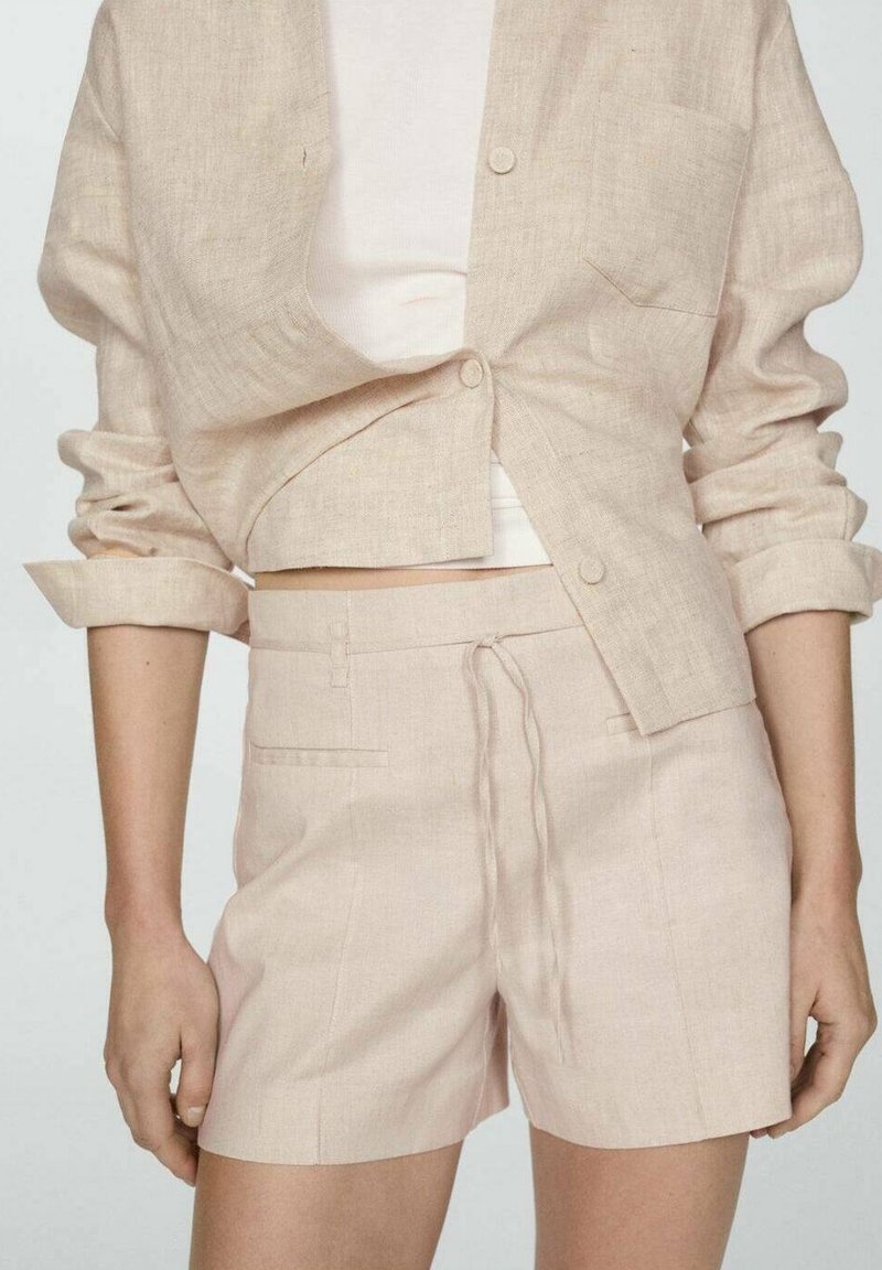 Veste beige en lin boutonnée avec manches retroussées et short assorti à cordon, portée par-dessus un top blanc ajusté.